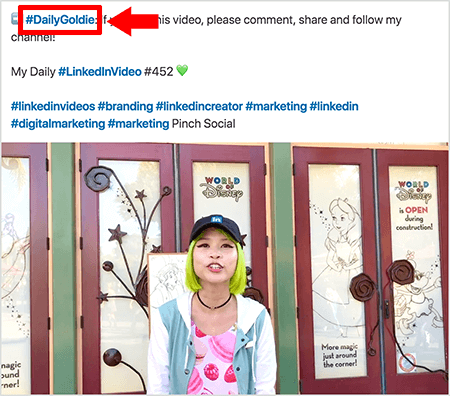 Esta é uma captura de tela que ilustra como Goldie Chan usa hashtags no texto de suas postagens de vídeo no LinkedIn. Os textos destacados em vermelho apontam para a hashtag #DailyGoldie no texto, que é exclusiva para suas postagens de vídeo e a ajuda a rastrear compartilhamentos. A postagem também inclui outras hashtags relevantes que ajudam as pessoas a encontrar seu vídeo, incluindo #LinkedInVideo. Na imagem do vídeo, Goldie está na frente de algumas portas em uma exibição do World of Disney. Ela é uma mulher asiática com cabelo verde. Ela está usando um boné preto do LinkedIn, uma gargantilha preta, uma camisa rosa com estampa macaron e uma jaqueta azul e branca.